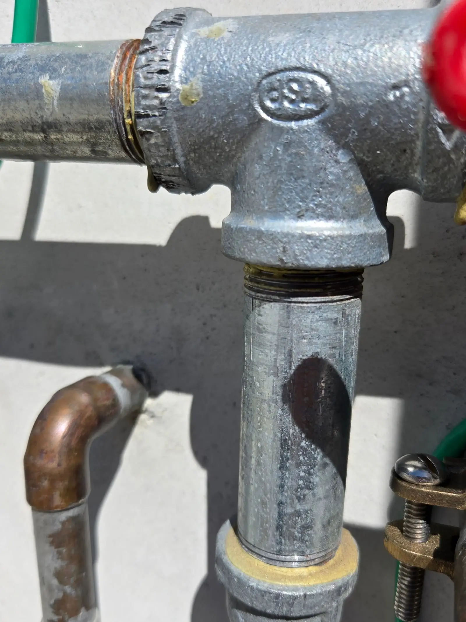 Close view of gray galvanized steel pipe joined by a threaded tee fitting, with a copper elbow below it and the edge of a red valve handle at the right