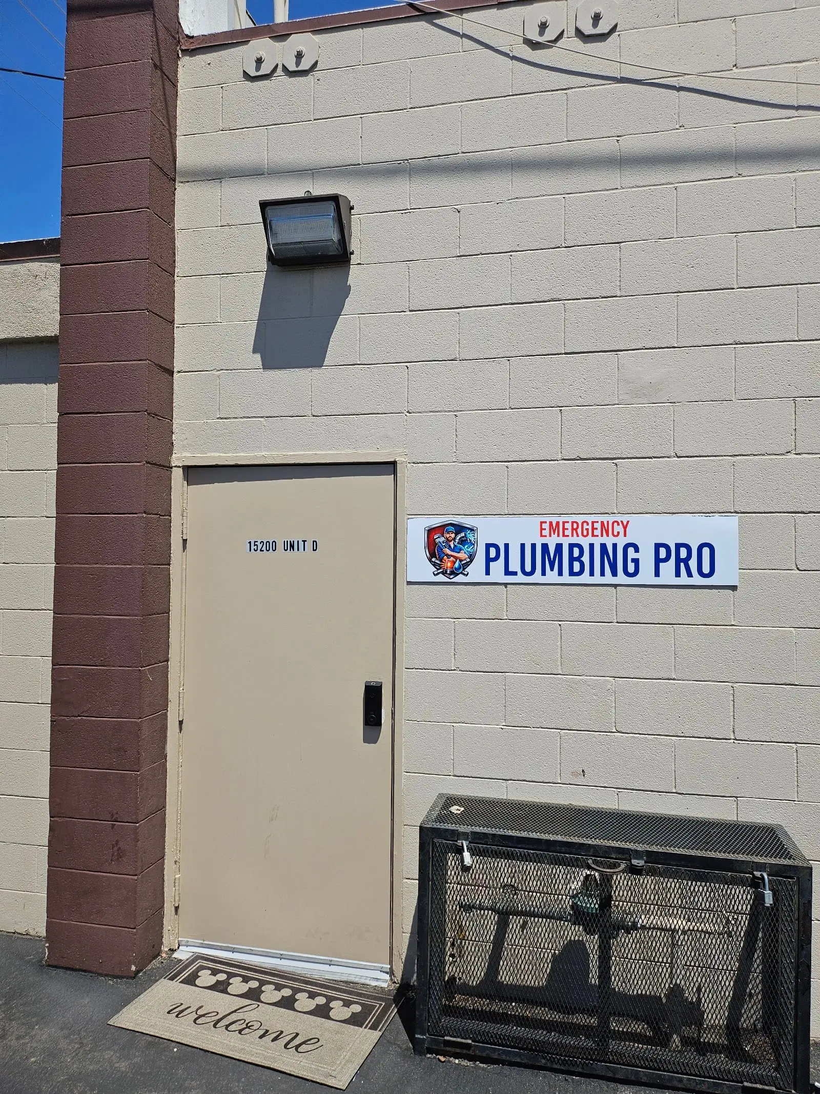 Beige concrete block wall with a tan door marked 15200 Unit D, an Emergency Plumbing Pro sign beside it, a wall light above and a black metal cage with pipework inside on the ground at the right