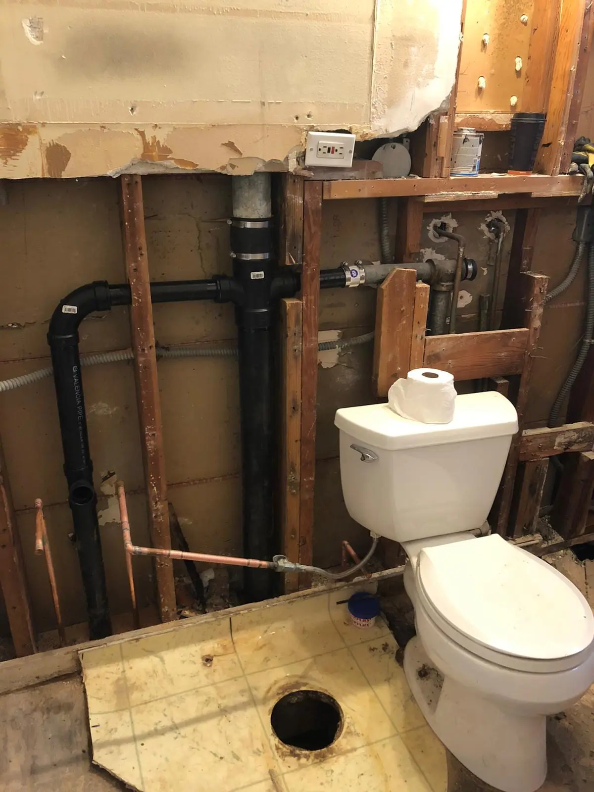 Bathroom wall opened to the studs, showing a black drain stack with branch lines, copper supply pipes and a white toilet beside a hole cut in the floor